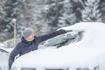 A man in a dark jacket and gray hat diligently removes a thick layer of fresh snow from the windshield of his car with a scraper and a broom. 