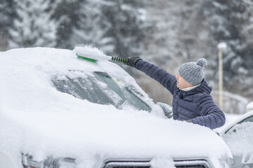 A man in a dark jacket and gray hat diligently removes a thick layer of fresh snow from the windshield of his car with a scraper and a broom. 