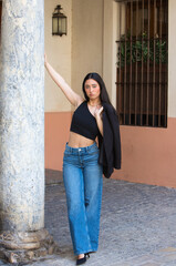 A young, attractive woman with dark hair, dressed in jeans and a black top holds her jacket in her hand over her shoulder and leans against a marble column on a street in the centre of Seville, spain.