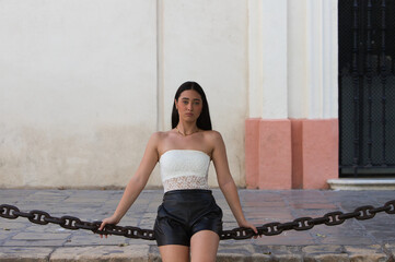 A young, attractive Spanish woman dressed in a white lace top, black shorts and black leather boots, sitting as if on a swing on a chain attached to two stone columns.