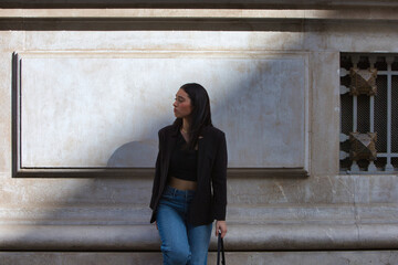 A young, attractive Spanish woman dressed in casual clothes and carrying a black handbag looks serious and worried as she leans against the wall of a building and looks from side to side.