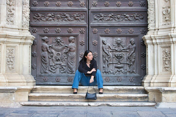 A young, attractive Spanish woman dressed in casual clothes and carrying a black handbag sits casually but seriously on the steps of the Plateresque-style entrance to Seville City Hall.