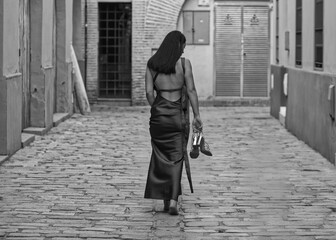 A young, dark-haired woman wearing a brown dress and holding gold heels in her hand, with her back turned, walks barefoot down a lonely alleyway. Her feet plant firmly on the ground. Monochrome photo.