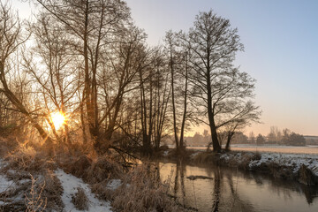 Winter sunrise over a small river in rural Poland, with frost-covered trees, calm water reflections, and a peaceful natural landscape. Cold morning light illuminates the snowy riverbank and bare branc