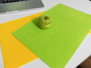 Green and yellow felt sheets with a ball of thread on a white desk. Creative DIY craft workspace with laptop flat lay