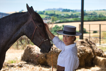 A young, beautiful woman dressed in riding clothes and a traditional Spanish hat is standing next to her grey horse in the barn. The woman is in love with her horse and touches it.