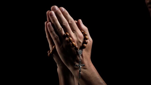 Close-up of hands clasped in prayer holding a wooden rosary. Person praying with beads and crucifix against a black background. Religious faith and devotion concept