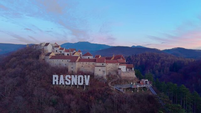 Aerial drone footage of Rasnov Fortress in Romania during a winter sunset. The camera pulls back from the fortress to reveal the surrounding mountain landscape, forested hills, and layered sky in the 