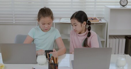 Two diverse elementary schoolgirls studying with laptops at home, one showing her Asian friend content on a smartphone for collaborative e-learning and modern education in this 4k real time footage. - Powered by Adobe