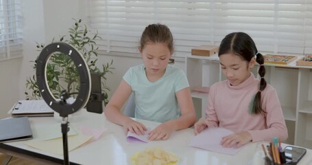 Two young diverse girl vloggers record a DIY origami tutorial using a smartphone and ring light at home, creating educational content for their online channel in this cinematic 4k real time clip.