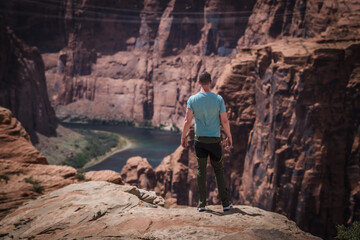 View of a man in a blue shirt gazes out over the precipitous red rock cliffs and the winding river below, bathed in sunlight, Page, Arizona, United States.
