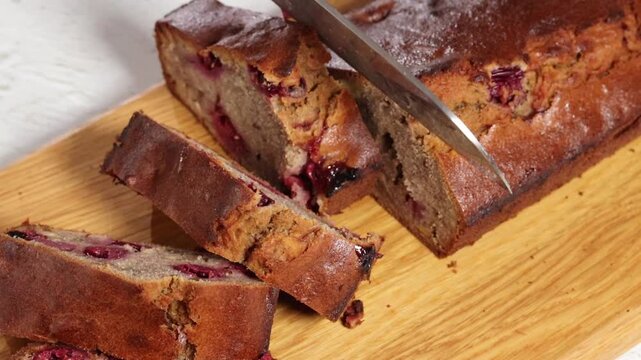 Banana bread. Slicing freshly baked cherry banana bread loaf cake on a wooden cutting board in a bright kitchen. The cake shows pieces of cherries inside and has a golden crust.