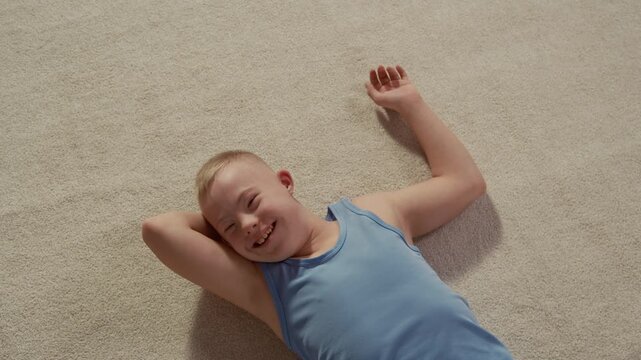 Caucasian boy with down syndrome wearing blue undershirt resting on rug and smiling at camera, conveying positivity and joy