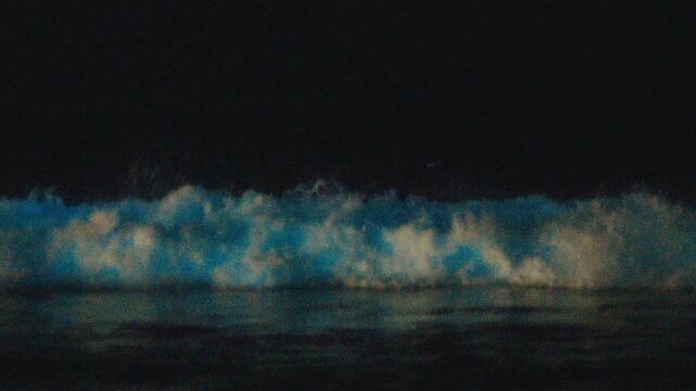 Bioluminescence of the ocean wave. Night view of the sea surface with breaking wave shining strong blue light caused by bioluminescence. High level of noise