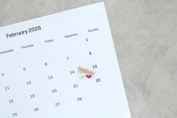 Calendar February 2026 showing Valentine's Day with red heart, small shell, and rolled paper scroll. Romantic planning and date reminder concept for holiday celebration on gray background.