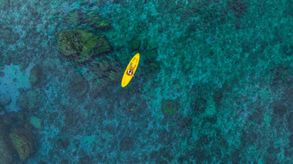 Aerial top view of a yellow kayak rowing in clear blue ocean water with underwater rocks and coral...