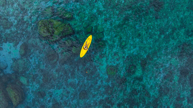 Aerial top view of a yellow kayak rowing in clear blue ocean water with underwater rocks and coral reefs, summer vacation and tropical sea adventure concept.