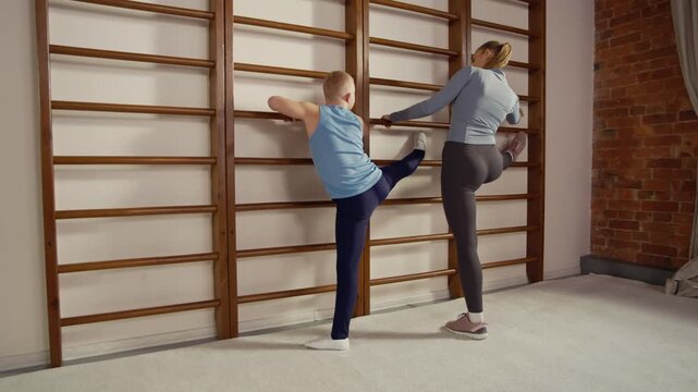 Young Caucasian oy with down syndrome stretching legs on wall bar with female fitness trainer in sports class