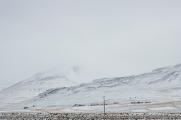 View of snow-covered mountains under a pale sky, contrasting with the muted tones of the landscape below, creating a serene yet stark scene, Blonduos, Vatnsnes peninsula, North Iceland.