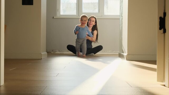 a child toddler with bare legs runs away from mother's arms across the floor against a sunny window, hugging mom happy family first steps towards his family at the window of the house