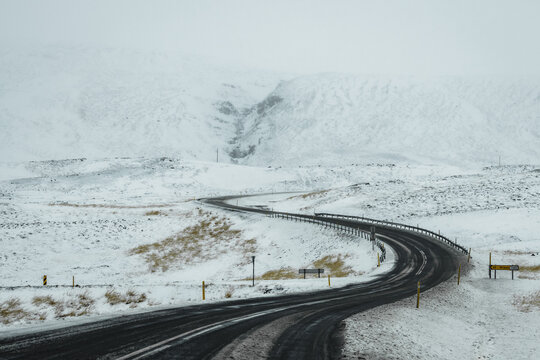 View of an asphalt road winds through a snow-covered landscape, cutting a dark path against the pristine white fields, Blonduos, Vatnsnes peninsula, North Iceland, Iceland.