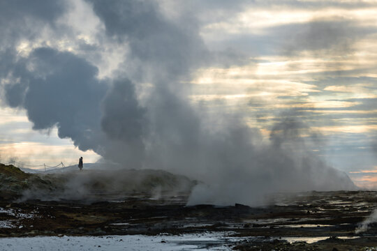 View of geothermal steam rising from the dark, textured earth, a solitary figure amidst the otherworldly landscape, under a sky streaked with dawn, Rejkjahlid, North Iceland, Iceland.