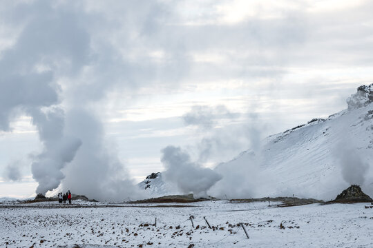View of geothermal steam rising from the stark, snow-dusted earth, contrasting with the distant figures in red, in Rejkjahlid, North Iceland, Iceland.