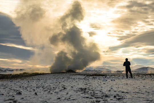 View of geothermal steam rising from the stark, snow-dusted earth as a lone figure gazes upon the surreal landscape, Rejkjahlid, North Iceland, Iceland.