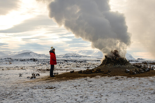 View of a person in a vibrant red jacket gazing at a steaming fumarole against a backdrop of snowy mountains and a cloudy sky, Rejkjahlid, North Iceland, Iceland.