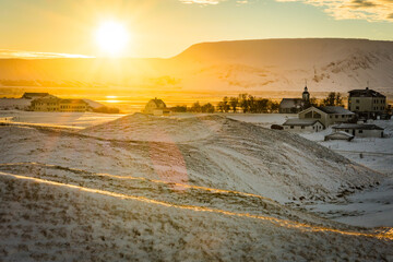 View of golden sunlight bathing a snow-dusted landscape, warming the quaint buildings of a village nestled beneath distant, snow-capped mountains, Rejkjahlid, North Iceland, Iceland.