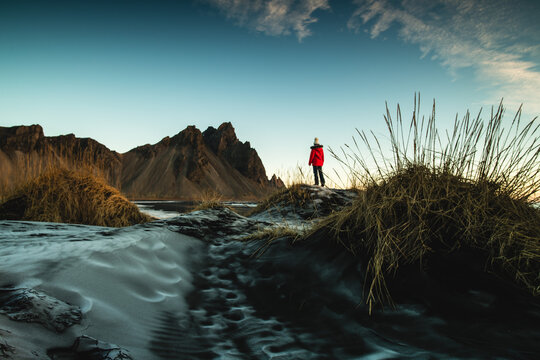 View of a person in a vibrant red jacket standing on a dune overlooking the majestic, dark Vestrahorn mountains against a clear sky, Hofn, Eastfjords region, Iceland.