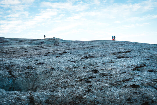 View of a distant figures standing on a glacier under a pale sky, the ice a mosaic of blues and grays, etched by time and elements, Vatnajokull, Iceland.