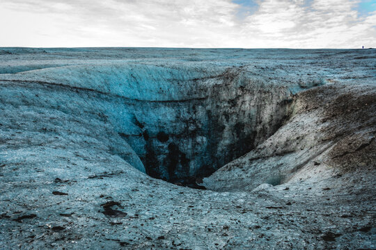 View of a sapphire ice cave's gaping maw reveals a dark abyss within the glacier's icy expanse, illuminated by ethereal blue hues, Vatnajokull, Iceland.