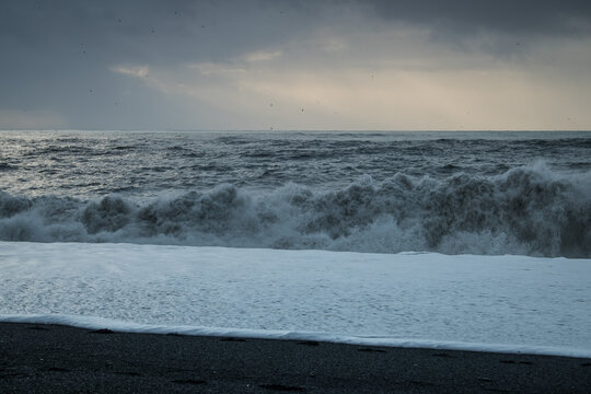 View of dark, turbulent waves crash onto the black sand beach under a moody sky, creating a stark contrast between the sea and shore, Vik, Iceland.