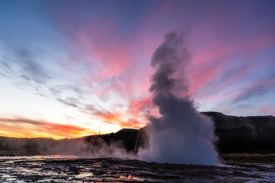 View of Geysir erupts in a burst of scalding water against a backdrop of fiery sunset hues painting the Icelandic sky, Haukadalur valley, Southwestern Iceland, Iceland.