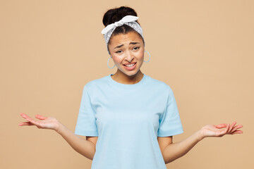 Young sad woman of African American ethnicity wear blue t-shirt headscarf casual clothes shrugging shoulders looking puzzled spread hands isolated on plain beige background studio. Lifestyle concept.