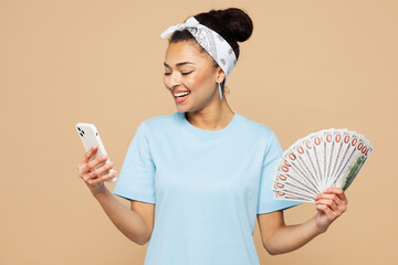 Young happy woman wear blue t-shirt casual clothes hold in hand fan of cash money in dollar banknotes use mobile cell phone isolated on plain pastel light beige background studio. Lifestyle concept.