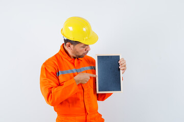 Young worker in uniform, helmet pointing at blackboard and looking focused , front view.