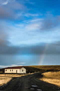 View of a lonely house with a red roof stands beside a gravel road under a blue sky with clouds and a faint rainbow, Southern Iceland, Iceland.