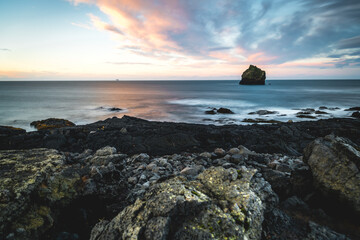 View of the rugged, dark volcanic rocks meeting the serene, blurred ocean under a pastel sky with a solitary rock formation, Keflavik, Iceland.