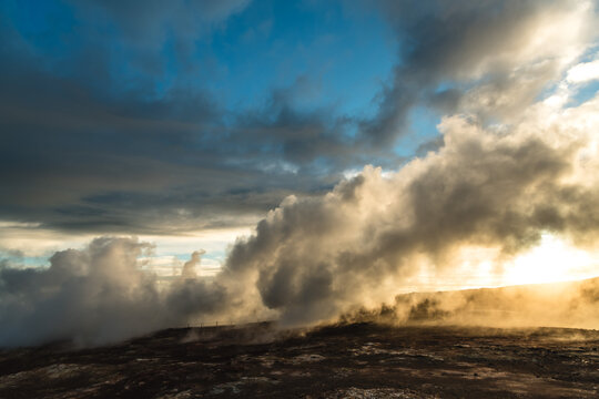 View of geothermal steam rises dramatically from the dark, rugged terrain under a sky streaked with blues and grays, Hveragerdi Geothermal Park, Keflavik, Iceland.