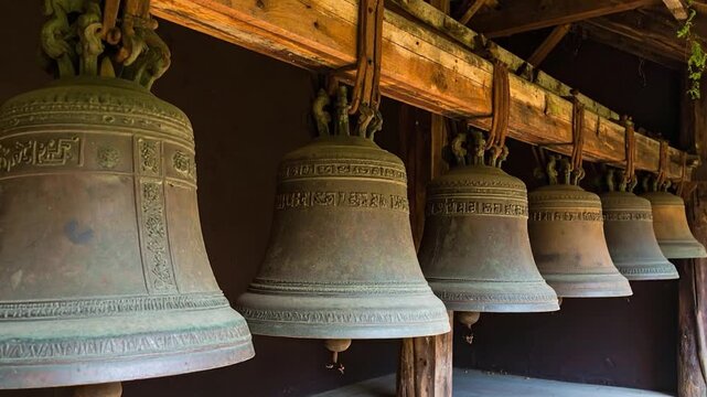 A row of aged bronze bells, each suspended, hangs in a wooden shelter; light highlights the textures