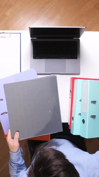 Vertical video person arranging multiple ring binders and documents on a white desk next to a laptop, fostering organization, productivity, and office management
