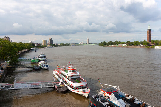 High-angle panoramic view of the Rhine river in Cologne, Germany, with several tourist excursion boats docked at the pier and a city skyline under a cloudy sky