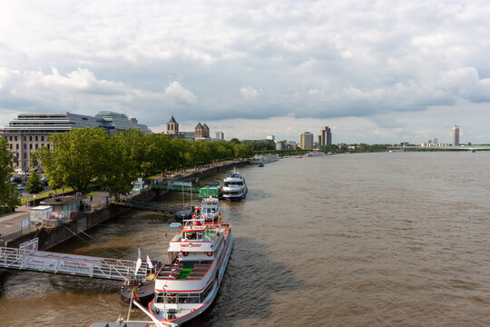 High-angle panoramic view of the Rhine river in Cologne, Germany, with several tourist excursion boats docked at the pier and a city skyline under a cloudy sky