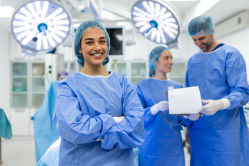 Portrait of a confident smiling female surgeon standing with arms crossed in a modern operating room, with colleagues in blue scrubs and surgical lights in the background of a hospital clinic.