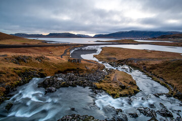 View of cascading waters flow over jagged rocks into a tranquil bay, framed by rugged hills under a dramatic sky, Grundarfjordur, Iceland.