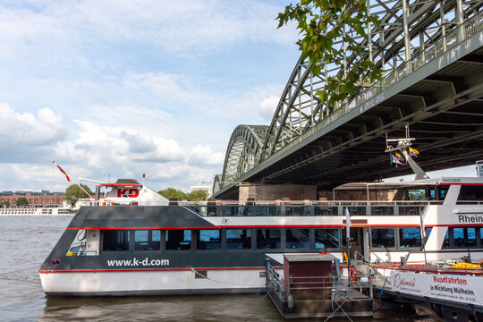 A passenger sightseeing boat docked on the Rhine river next to the steel arches of the Hohenzollern Bridge in Cologne, Germany