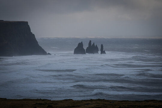View of jagged sea stacks rise defiantly from the turbulent, white-capped ocean under a brooding sky, a stark contrast to the dark cliffs, Vik, South Island, Iceland.