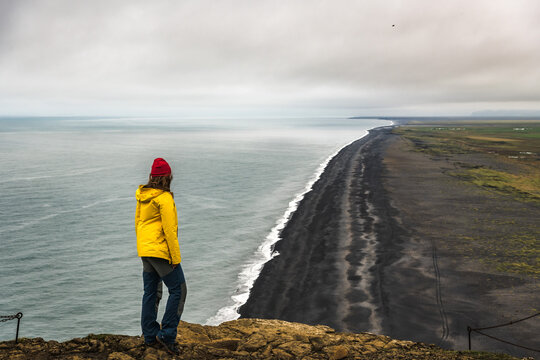 View of a person in a yellow jacket and red hat standing on a cliff overlooking the black sand beach and ocean under a cloudy sky, Vik, Iceland.